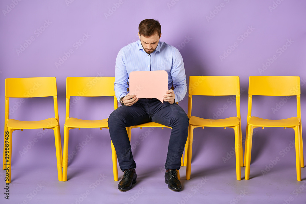 Young man sitting on yellow chair looking at blank thought bubble and thinking waiting for job interview Young man sitting on yellow chair looking at blank thought bubble and thinking waiting for job interview