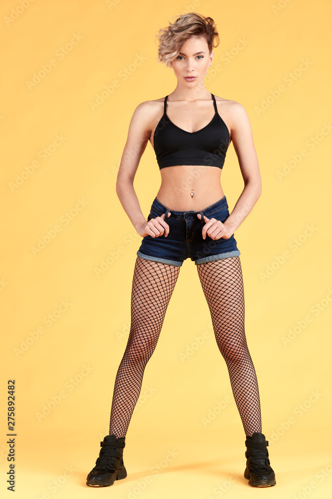 exciting total Perceive Female wearing black top, shorts, fishnet pantyhose, black trendy shoes  standing with hands on her hips, isolated on yellow.full length photo.  Stock Photo | Adobe Stock