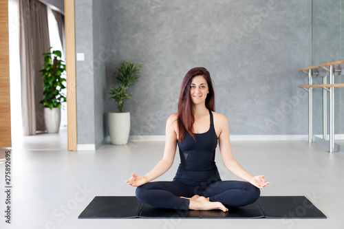 Young woman practices yoga. Girl is smiling and sitting on mat indoors. Sport workout in gym. Concept of calmness, relax, healthy lifestyle, wellbeing. Female fitness classes.