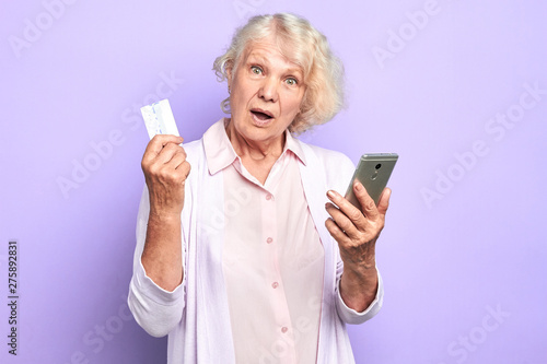 old doctor with surprised expression holding mobile phone and credit card. old woman cannot understand how she hasspend money, lost money. watch out, mobile fraudsters. isolated light blue background.