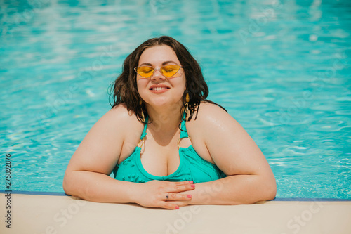 Attractive busty curvy woman in yellow sunglasses and a blue swimsuit resting by the pool. Stylish accessories, fringe, fashion for plus size. Bodypositive, natural real beauty, resort, summer