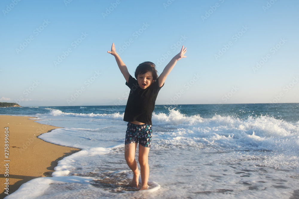 Boy in black t-shirt have fun by the sea. Child play on coastline while big wave splashing on beach