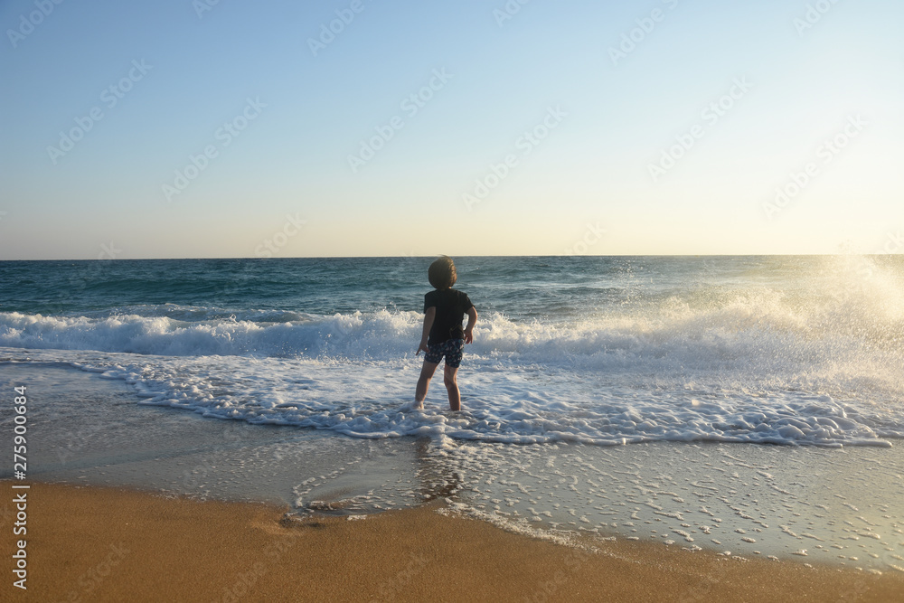 Boy in black t-shirt have fun by the sea. Child play on coastline while big wave splashing on beach