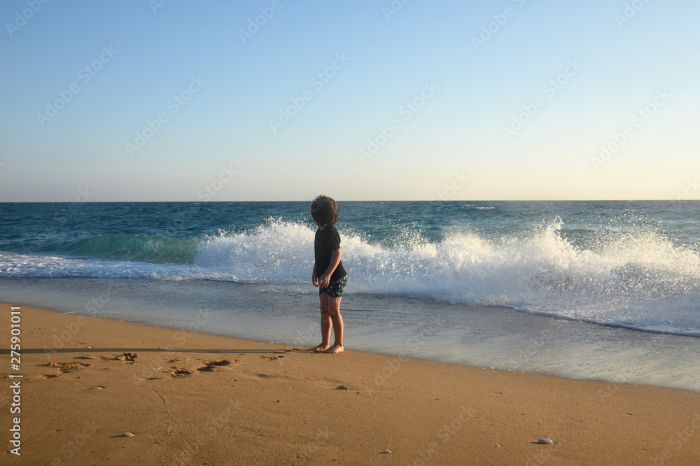Boy in black t-shirt have fun by the sea. Child play on coastline while big wave splashing on beach