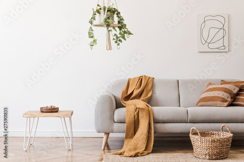 Modern boho interior of living room at cozy apartment with gray sofa, honey yellow pillows and plaid, plants, paintings, rattan basket and design personal accessories. Stylish home decor. Template.