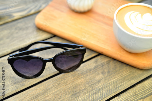 cup of coffee on a wooden stand is on the table in a cafe summer morning, sunglasses in the foreground