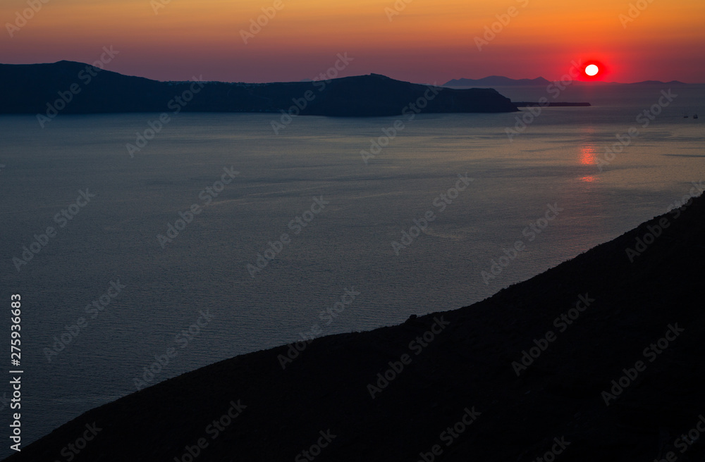 Fototapeta premium Oia santorini island greece. Natural coloured sunset. Dark red sky. Gold setting sun behind mountains. Calm sea reflecting sunlight. Beautiful colours. Thin clouds slightly visible.