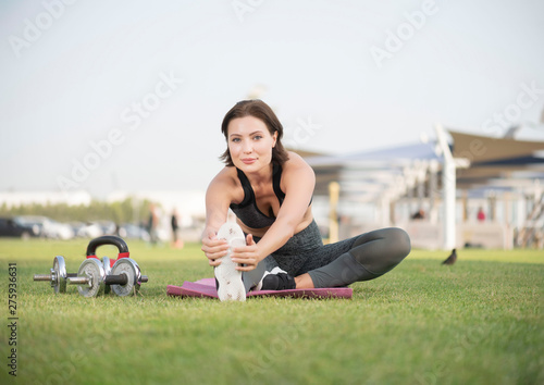 Beautiful caucasian brunette fitness model does a hamstring leg stretch on a pink yoga mat in a open park on a bright sunny day