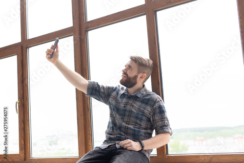 Side view of a positive handsome hipster guy and taking a selfie on a smartphone while sitting on a windowsill by a large window. Concept of gadgets and modern technologies.