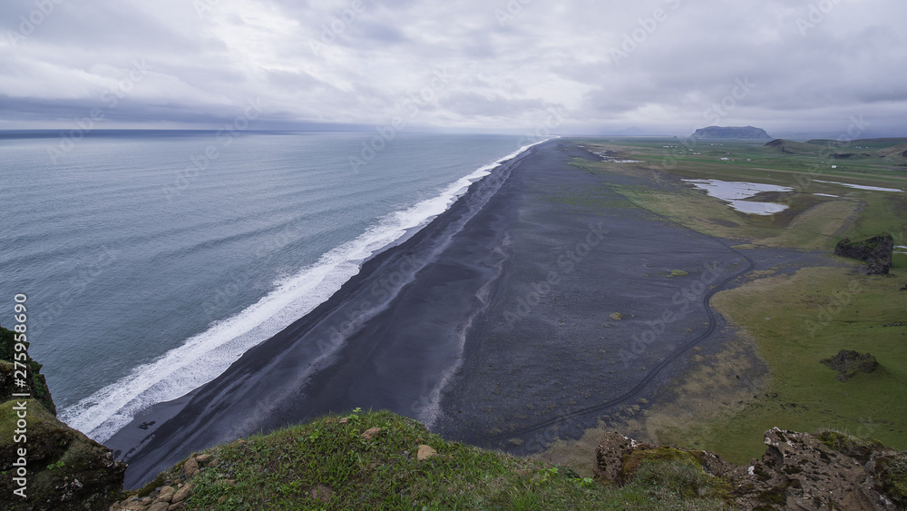 Iceland's Black Beach at Dyrholaey seen from high above. Surf line diagonal. Atlantic on left, black sand beach and grassy lowland right. An inselberg in distance. Iceland's Black Beach at Dyrholaey seen from high above. Surf line diagonal. Atlantic on left, black sand beach and grassy lowland right. An inselberg in distance.