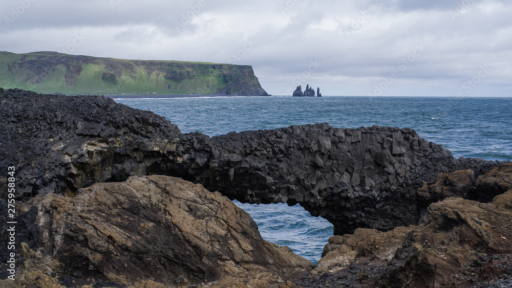 Summer on Iceland at Dyrholaey nature reserve -a look across a bay through a natural arch made by an eroded volcanic rock.  Ocean waves in centre,  mountains above coast with more sharp cliffs in back