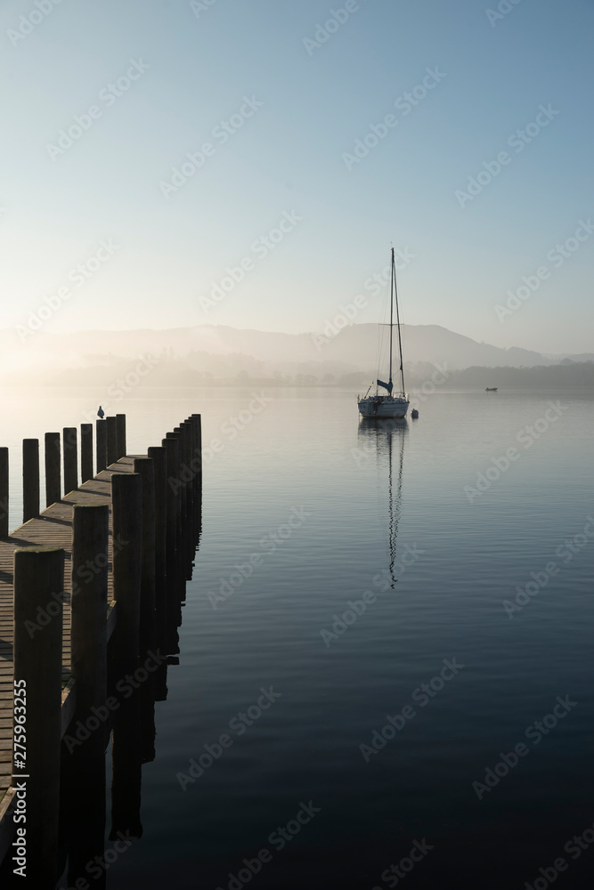 Obraz premium Stunning unplugged fine art landscape image of sailing yacht sitting still in calm lake water in Lake District during peaceful misty Autumn Fall sunrise