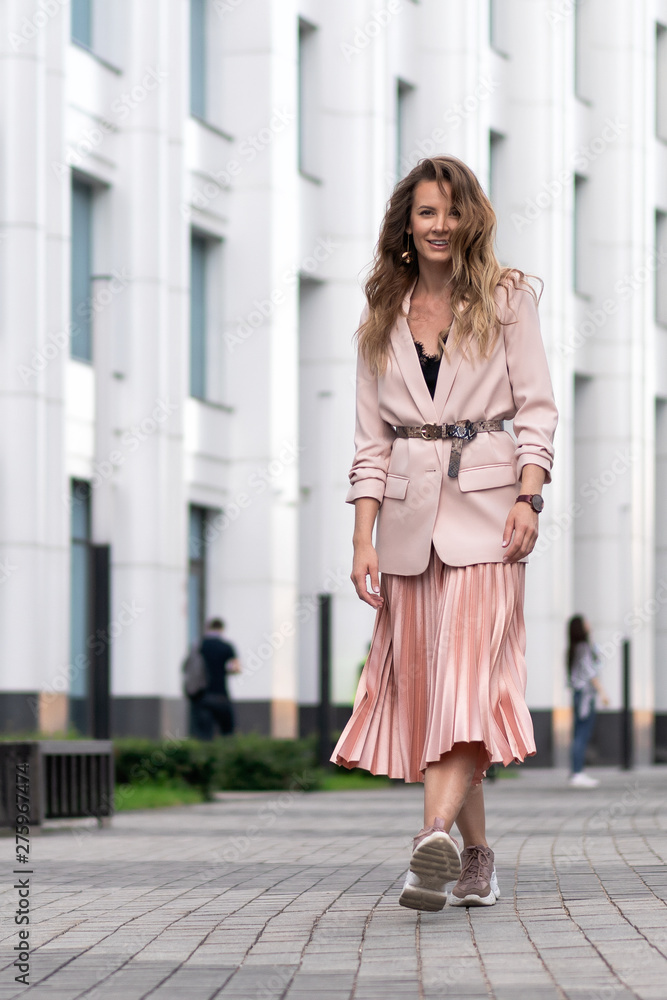 Women's clothing coral color. Fashionable look. Pink, peach color women's jacket, long skirt, pleated. The girl walks the street. Long wavy hair, pretty face, medium height. Copy space