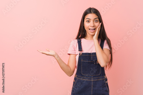 Young pretty arab woman wearing a jeans dungaree holds copy space on a palm, keep hand over cheek. Amazed and delighted. © Asier