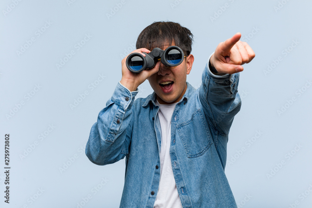Young chinese man holding a binoculars cheerful and smiling pointing to front Young chinese man holding a binoculars cheerful and smiling pointing to front