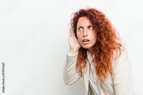 Young natural redhead business woman isolated against white background trying to listening a gossip.