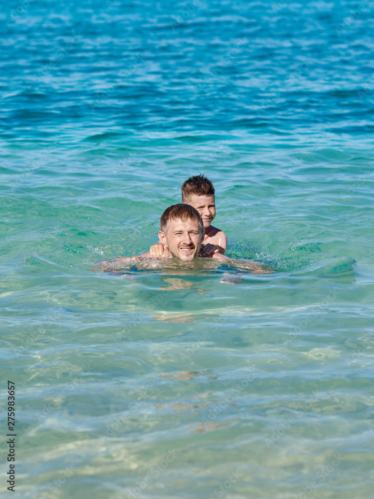 Dad and son are swimming in the ocean while on summer holidays. They are happy and smiling to the camera.