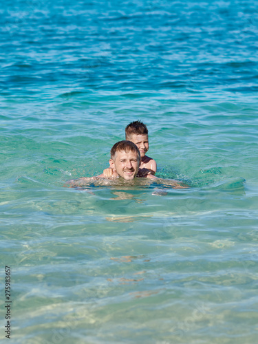 Dad and son are swimming in the ocean while on summer holidays. They are happy and smiling to the camera.