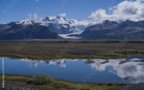 Polar summer in Iceland. Sunlit Mountains covered partly by snow reflect in a lake together with white clouds. A glacier visible across a patch of green between the mountain peaks. © Martin