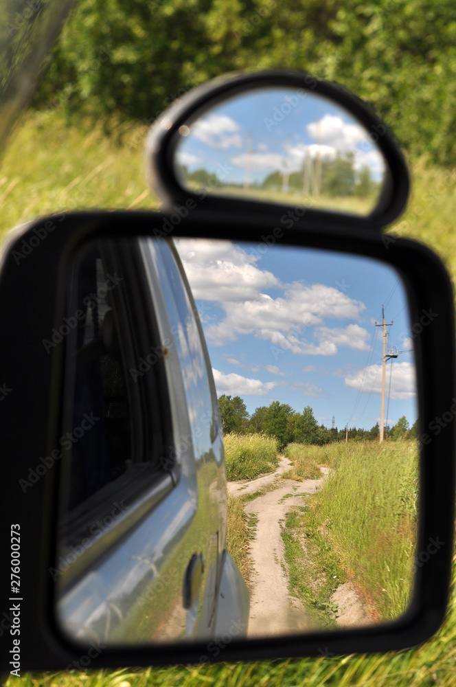 View in the side mirror of the car on a green meadow with a dirt road and a blue sky with clouds