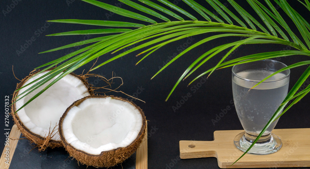 Exotic fruit Coconut, two halves of fruit in the shell, dark background, a glass of coconut water on the kitchen and blackboard, green palm trees, selective focus Exotic fruit Coconut, two halves of fruit in the shell, dark background, a glass of coconut water on the kitchen and blackboard, green palm trees, selective focus