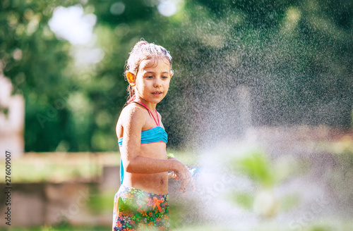 Cute little girl sprinkls a water for herself from the hose, makes a rain. pleasure for hot summer days. Happy careless childhood concept image. photo