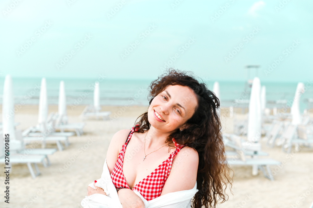 A woman in a red dress and curly hair is standing on the beach. A man with happy emotions. Walk along the sea promenade. Travel and tourism in the summer. Copy space