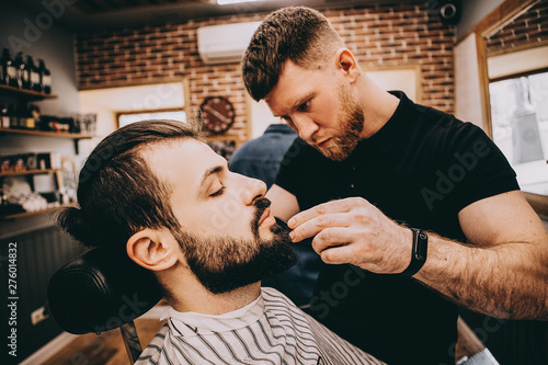 Getting perfect shape. Close-up side view of young stylish bearded man getting beard haircut by bearded, muscular hairdresser or barber at barbershop.Selective focus, noise effect