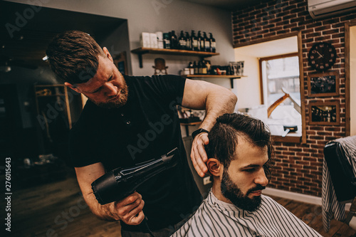 A stylish bearded client sits in a chair, a muscular barber cuts his black hair and makes a hairdo