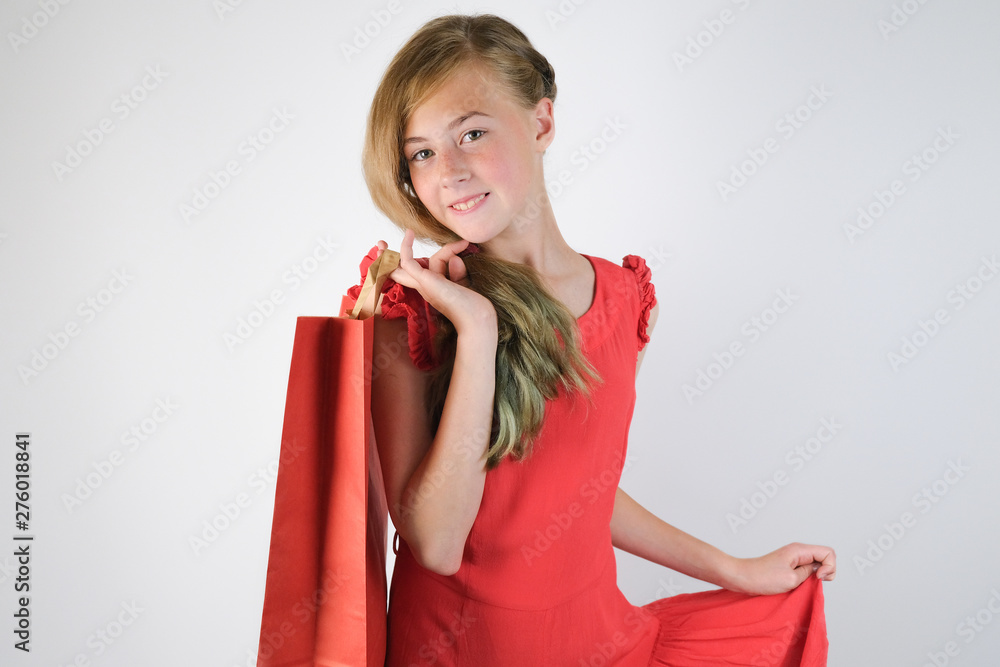 Attractive girl standing over grey background. Shopping concept. Beautiful blonde Caucasian girl in red dress holding a paper bag in her hand. Attractive girl standing over grey background. Shopping concept. Beautiful blonde Caucasian girl in red dress holding a paper bag in her hand.