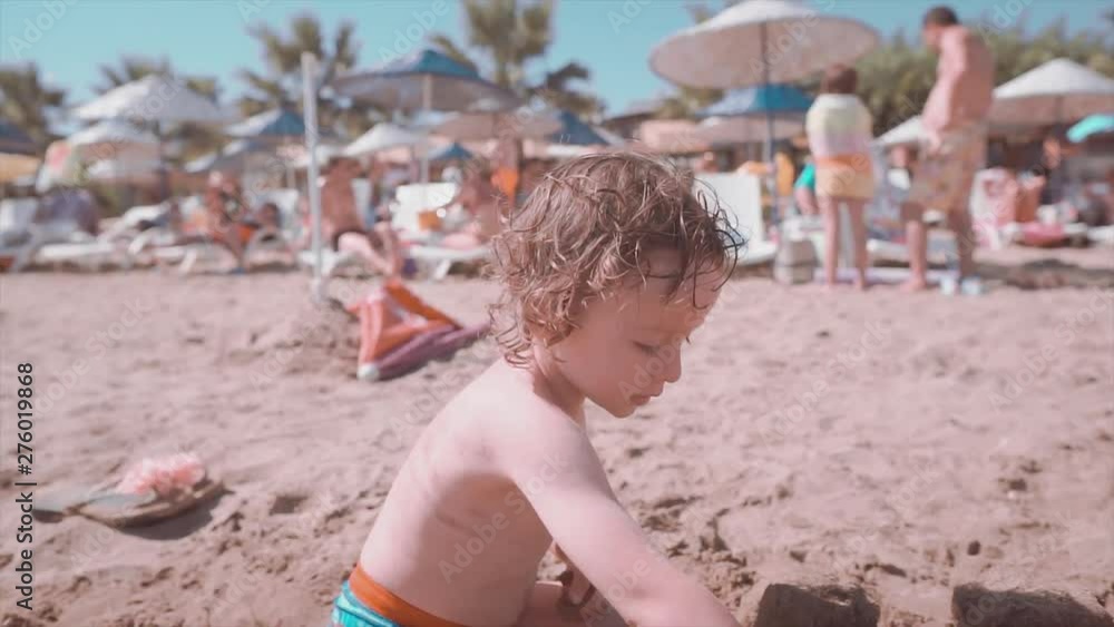 2 years old long haired boy is playing with sand on beach, summertime. Funny children making castle from sand, childhood and summer concept. 2 years old long haired boy is playing with sand on beach, summertime. Funny children making castle from sand, childhood and summer concept.