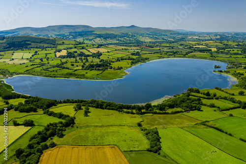 Fototapeta Naklejka Na Ścianę i Meble -  Aerial drone view of a lake surrounded by green farmland and fields in rural Wales (Llangorse Lake)