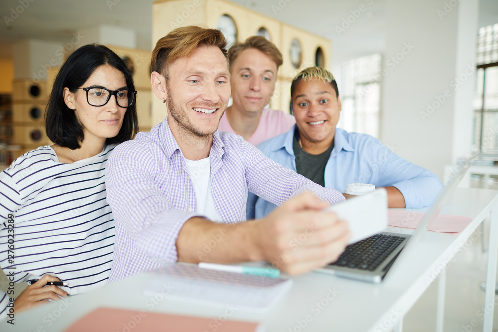 Group of cheerful hipster young multi-ethnic friends standing at counter with laptop and using gadget while photographing together in library Group of cheerful hipster young multi-ethnic friends standing at counter with laptop and using gadget while photographing together in library