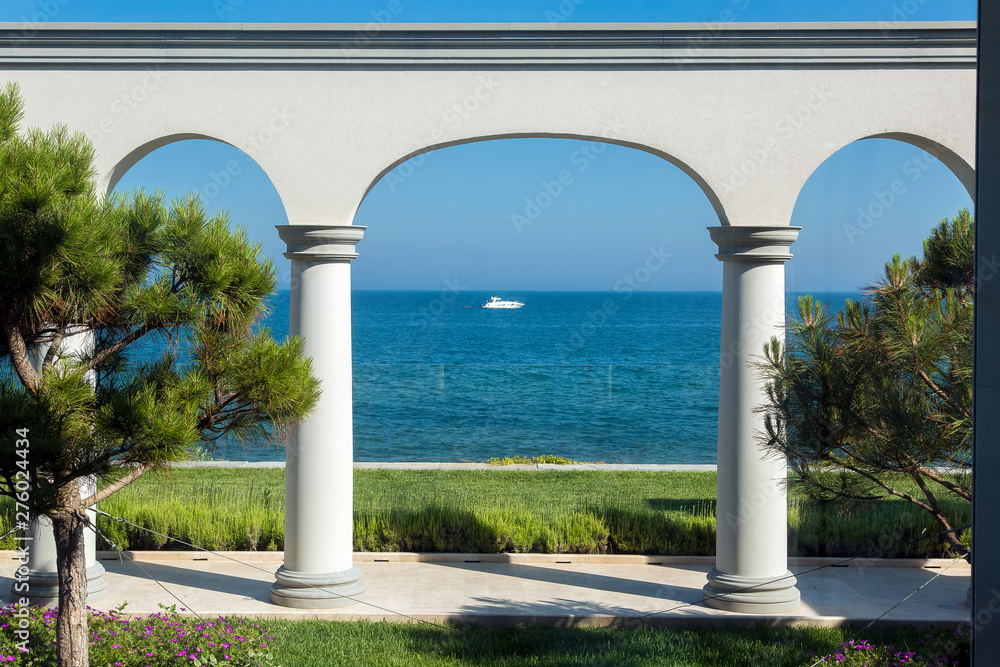 backyard with arched vaults and columns with a flowerbed of green lawns and pine trees in the background is a sea horizon with a yacht into the sea, tourism background.