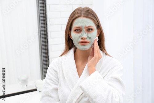 Beautiful woman removing homemade clay mask from her face indoors