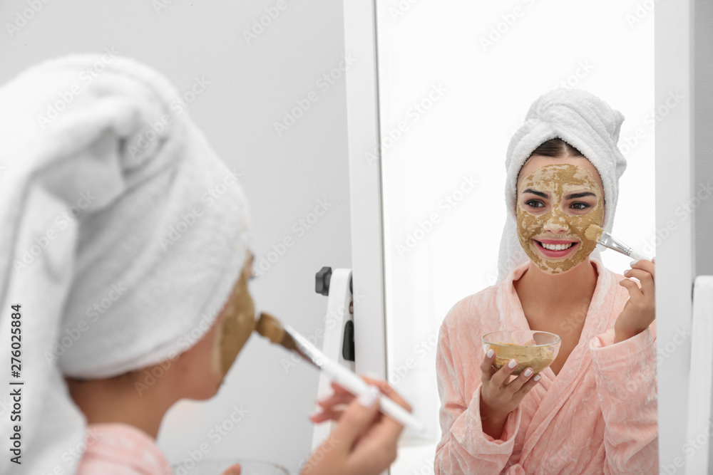 Young woman applying clay mask on her face near mirror in bathroom. Skin care Young woman applying clay mask on her face near mirror in bathroom. Skin care