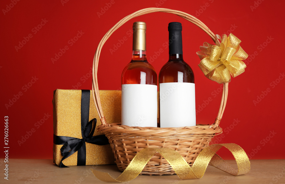 Bottles of wine in wicker basket with bow and gift on table against color background Bottles of wine in wicker basket with bow and gift on table against color background