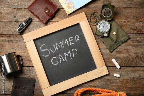 Chalkboard with text SUMMER CAMP and camping equipment on wooden background, flat lay