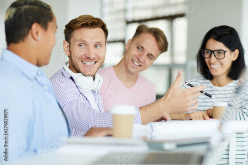 Positive enterprising young man with headphones on neck sitting at table and gesturing hand while explaining project idea to colleagues
