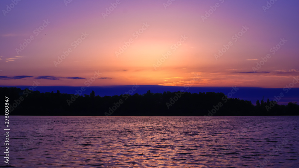 Very romantic sunrise over the water. Silhouette of an island in the distance with vegetation. Idyllic landscape with soft lights and colors in the sky.