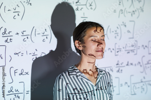Content calm young lady in blouse standing against projection screen with math formulas and keeping eyes closed at university class