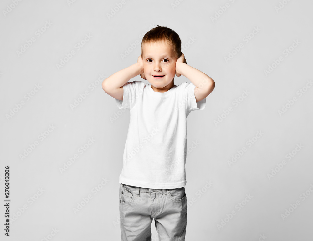 The little boy covered his ears with the palms of his hands. On a light background in a white T-shirt and gray pants The little boy covered his ears with the palms of his hands. On a light background in a white T-shirt and gray pants
