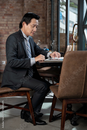 Mature Vietnamese entrepreneur in suit sitting at restaurant table and working with documents on tablet computer