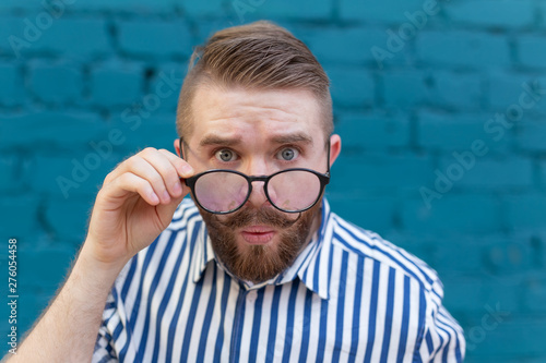 Billede på lærred Close-up portrait of a curious surprised young man in glasses with a mustache and beard posing on a background of blue blurred brick wall