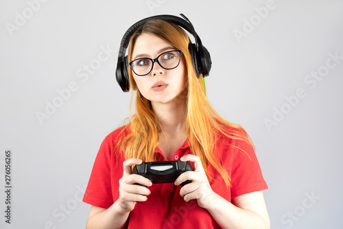 Beautiful young woman gamer in black headphones with a joystick in her hands on gray background. On girl black glasses and red shirt.