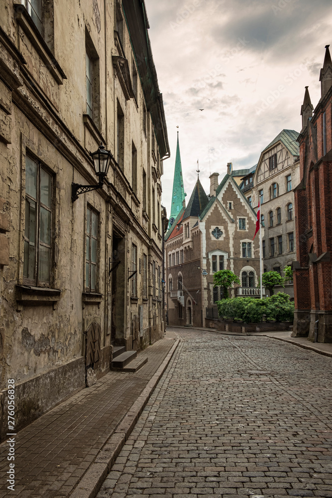 The narrow cobbled street of old Riga in  Latvia with not renovated dilapidated and shabby houses overlooking the spire of the Cathedral of St. James