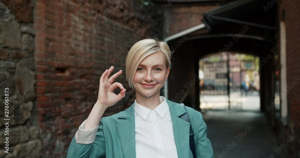 Slow motion portrait of attractive student girl showing OK gesture smiling outside in the street looking at camera. Happy youth, positivity and city concept. Slow motion portrait of attractive student girl showing OK gesture smiling outside in the street looking at camera. Happy youth, positivity and city concept.