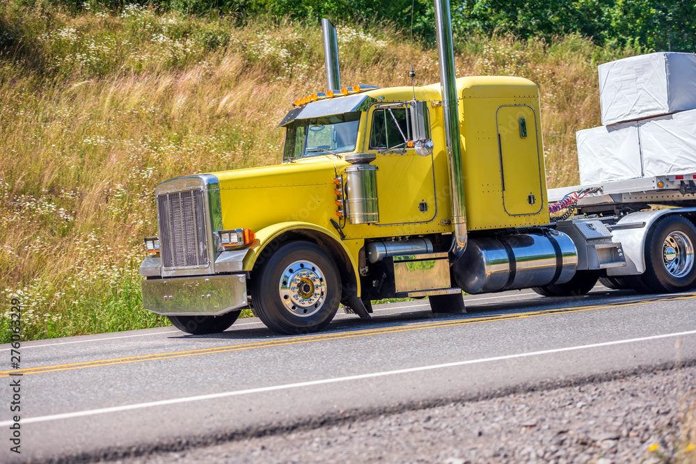 Classic yellow big rig semi truck transporting cargo on flat bed semi trailer running downhill Classic yellow big rig semi truck transporting cargo on flat bed semi trailer running downhill