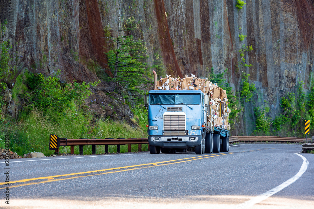 Old big rig cab-over semi truck transporting paper recycling on flat bed semi trailer driving on mountain winding road with rock wall