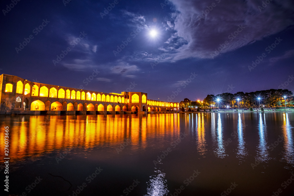 Khaju Bridge over Zayandeh river is iluminated at dusk with lights and moon in sky, Serving as a dam as well